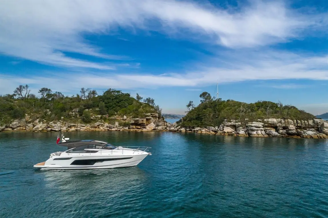A sleek white luxury yacht glides through calm waters near rocky shores and lush green hills under a clear blue sky.