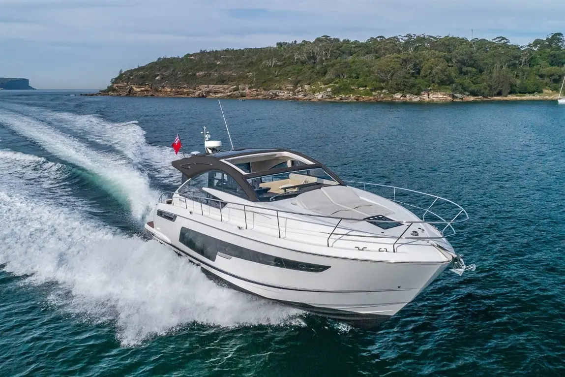 A luxury motor yacht speeds through clear blue water, leaving a wake behind; lush green hills are visible in the background.