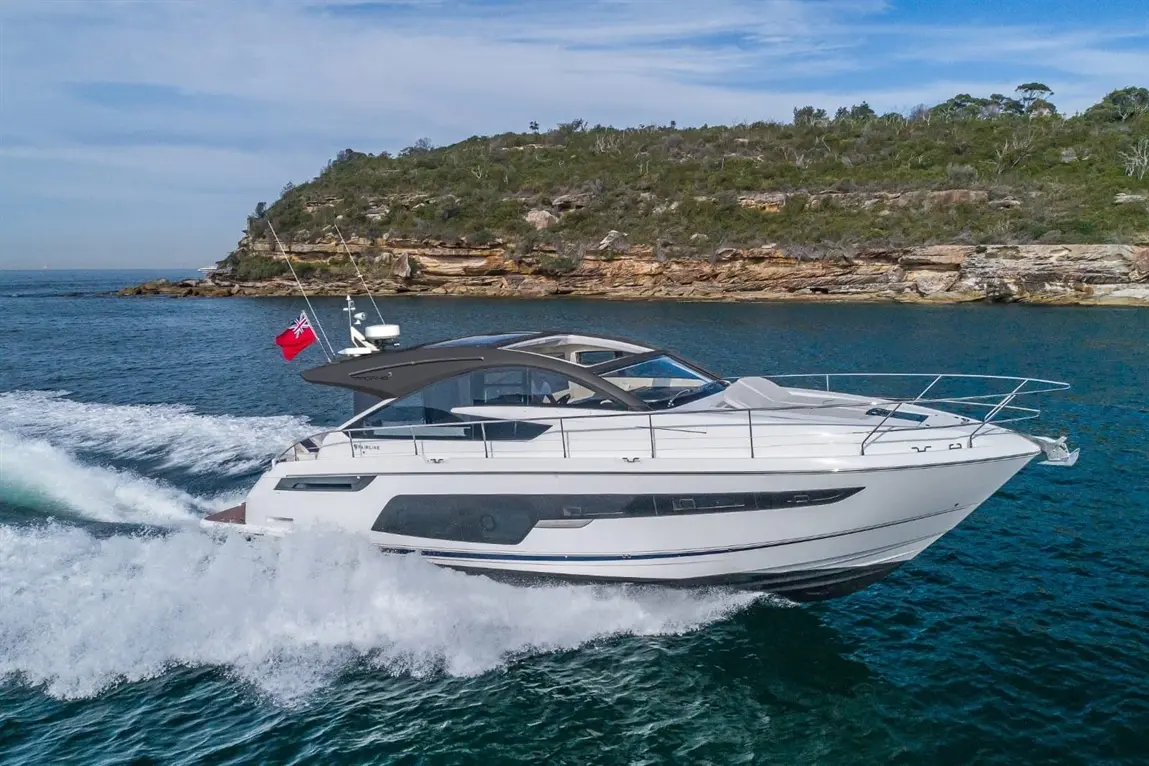 A sleek white motorboat speeds across clear blue water with a British flag flying, surrounded by rocky coastline and greenery.
