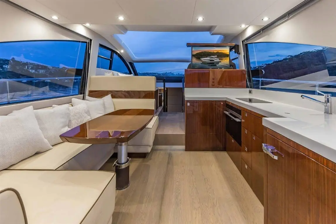 Modern boat interior featuring a dining area, kitchen cabinetry, plush cushions, and large windows showcasing a scenic view.