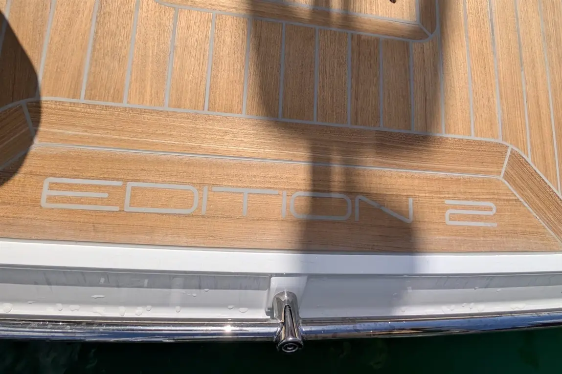 Wooden boat deck featuring the text "EDITION 2" in grey, illuminated by sunlight, with a sleek silver railing.