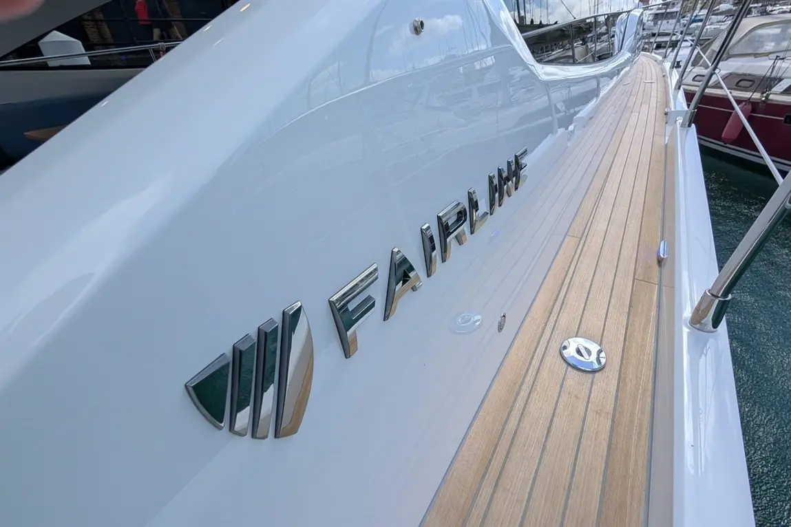 Close-up of a sleek white yacht with the "Fairline" logo and wooden decking, reflecting a marina in the background.