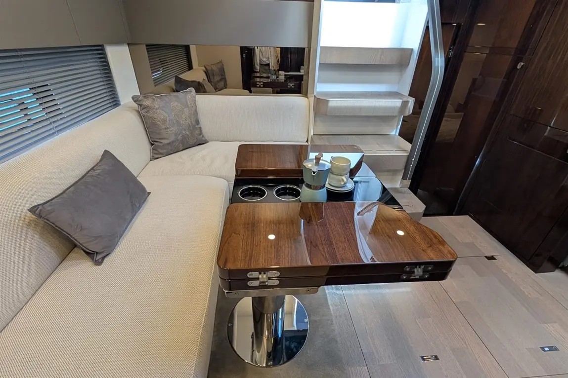 Stylish lounge area with a beige sofa, wooden coffee tables, a coffee pot, and a cup, alongside stairs in an elegant interior.