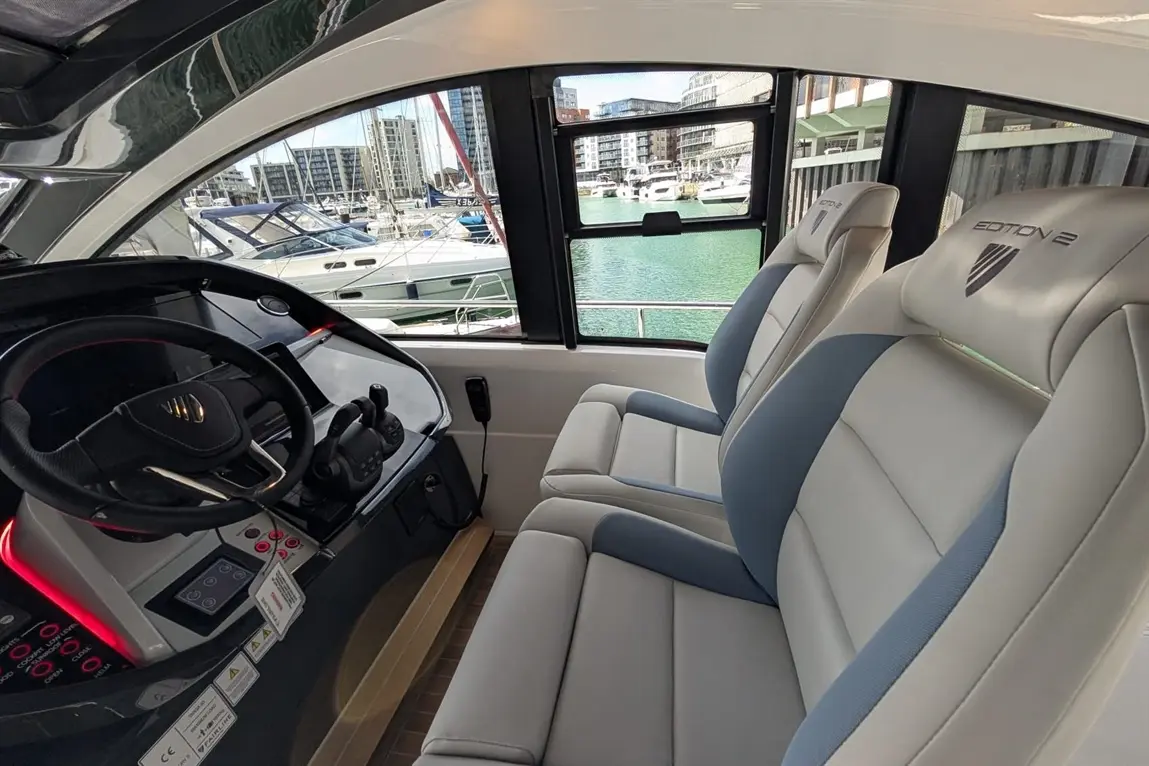 Interior view of a modern boat with two grey and blue seats labelled 'EDITION 2' and a control panel overlooking a marina.