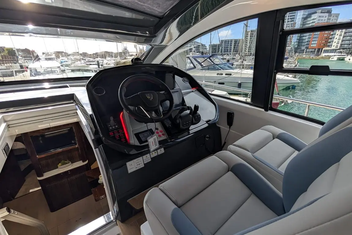 Interior of a modern boat's helm, featuring a steering wheel, controls, and two luxurious seats, with a marina view through windows.