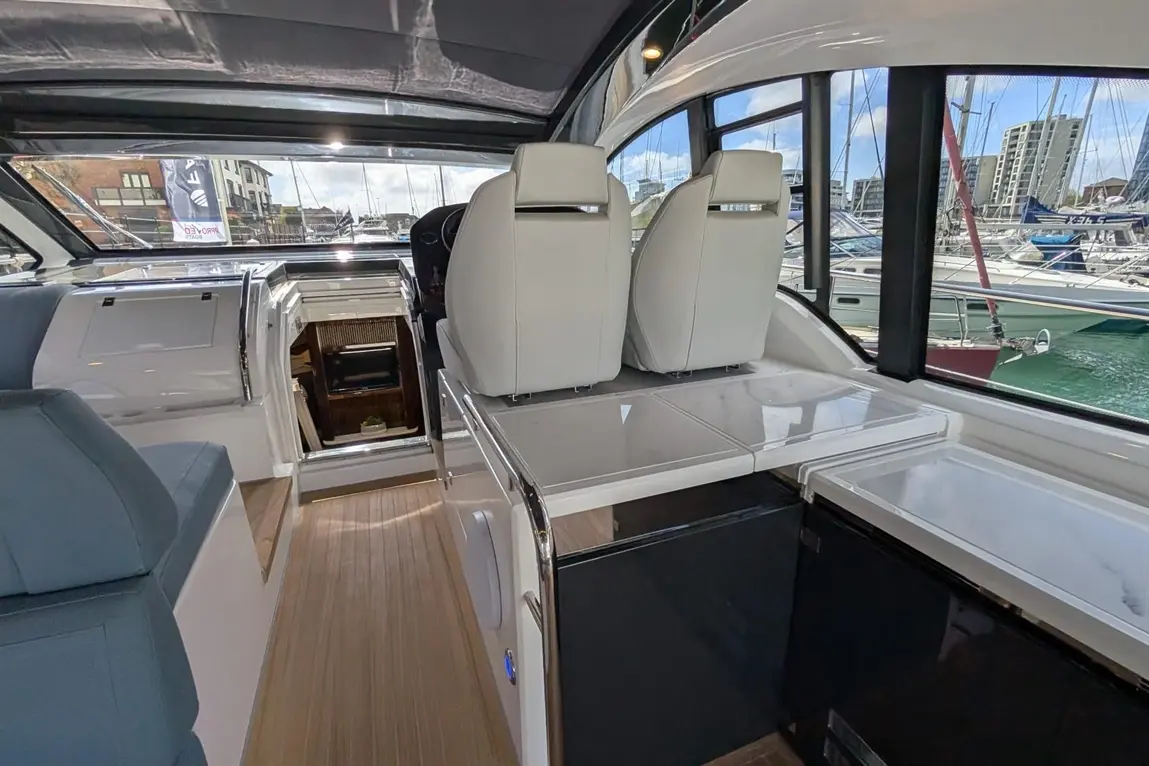 Interior of a modern boat with white seats, a cockpit area, polished surfaces, and marina views through large windows.