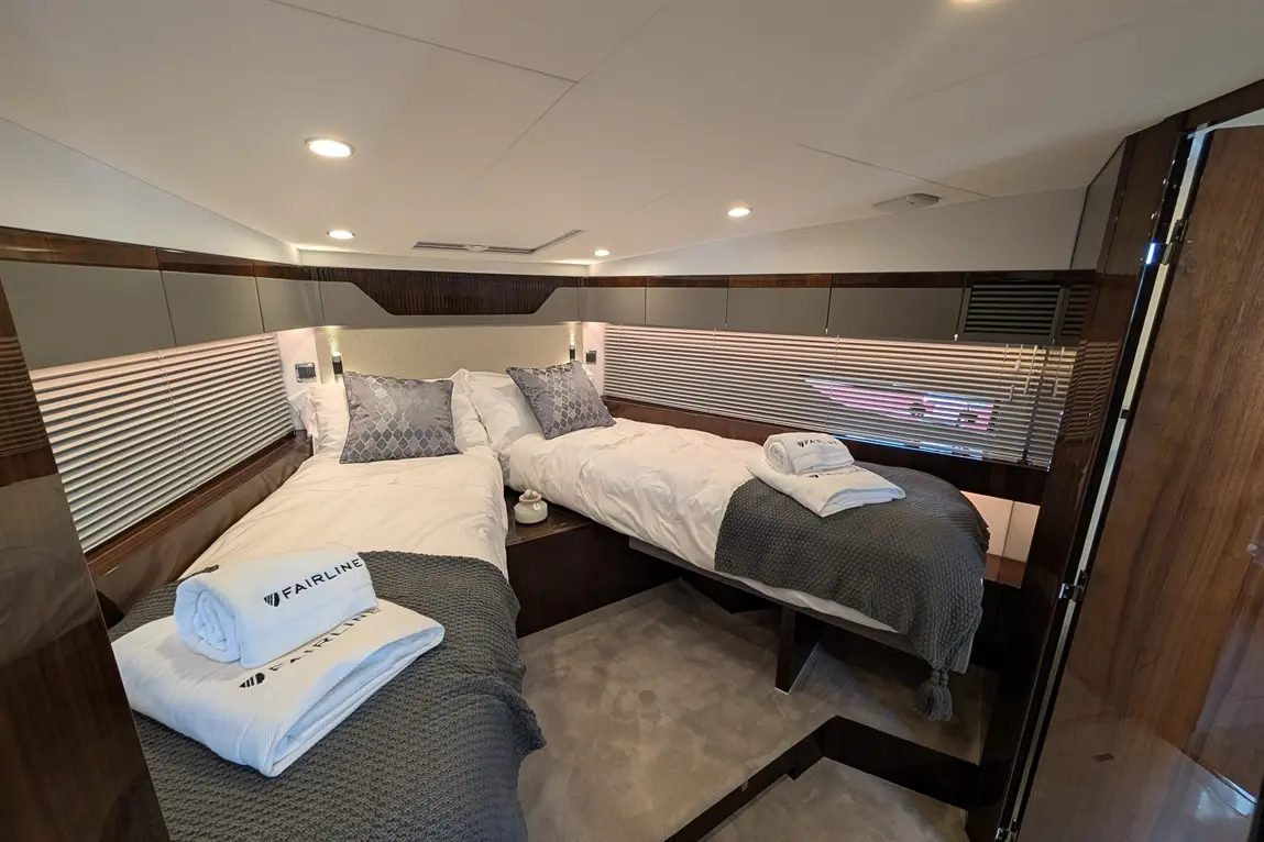 Modern yacht cabin featuring twin beds with grey throws, decorative pillows, and neatly folded towels labelled 'Fairline'.