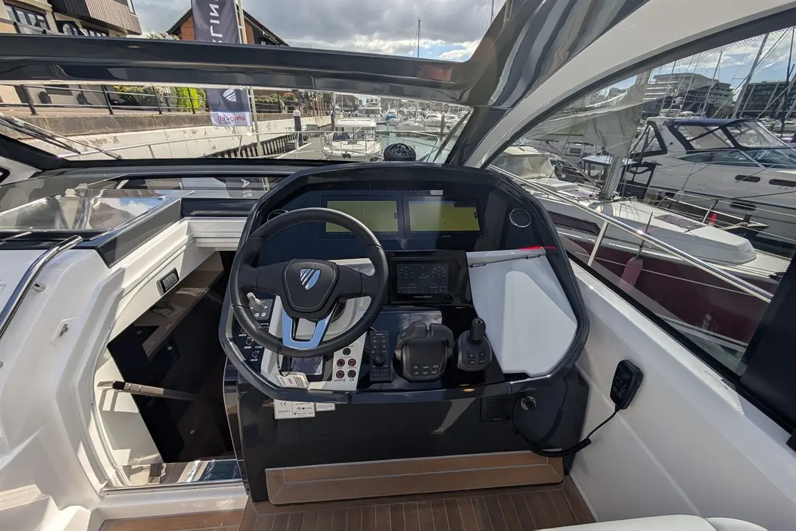 Luxury boat cockpit showing the steering wheel, control panels, and two screens, overlooking a marina with other boats.
