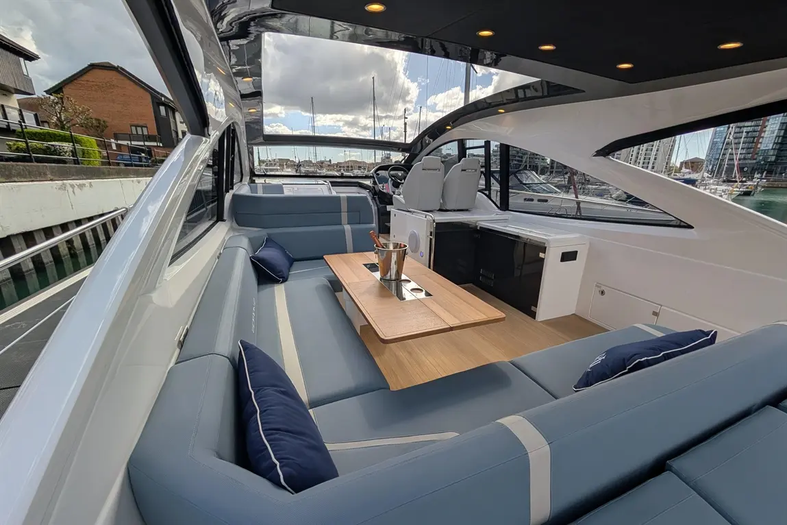Luxury yacht interior with comfortable seating, wooden table, and an ice bucket, overlooking a marina and cloudy sky.