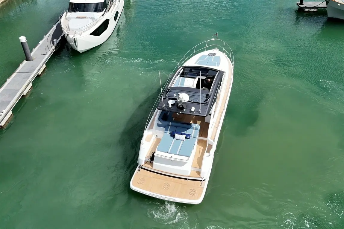 A sleek motorboat with a sunbathing area navigates bright green waters near a dock and another vessel in the background.
