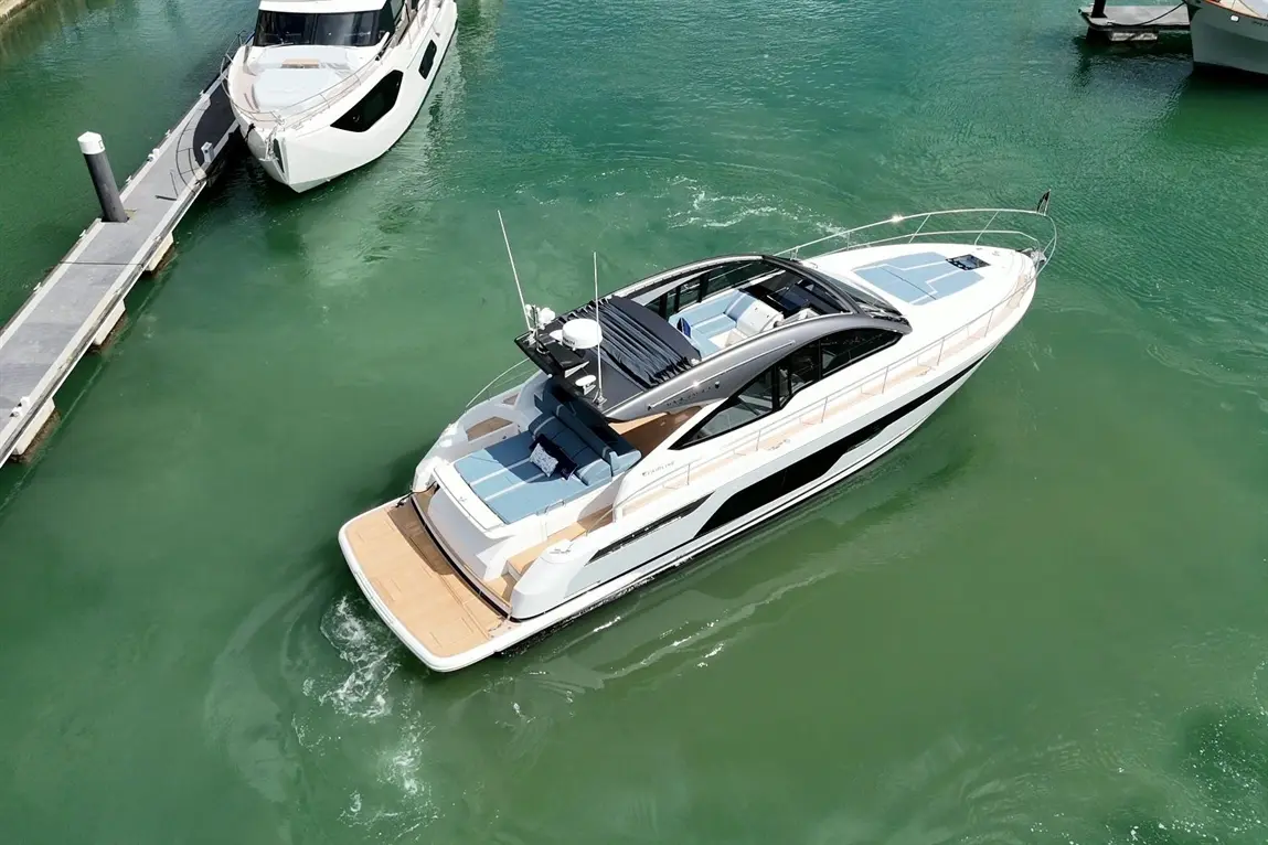 A luxury motor yacht with a sleek design navigates calm green waters near a wooden jetty and another boat in the background.