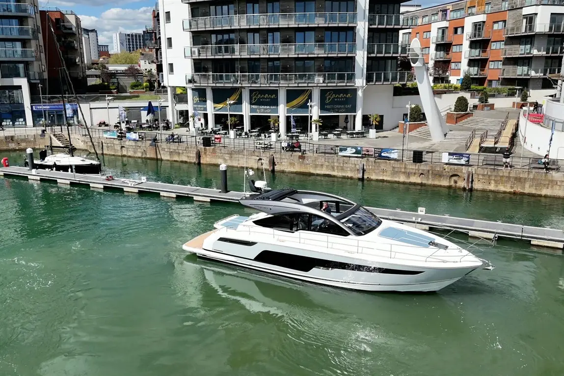 A luxury motor yacht navigates a marina with waterfront buildings and a café, under a clear blue sky.