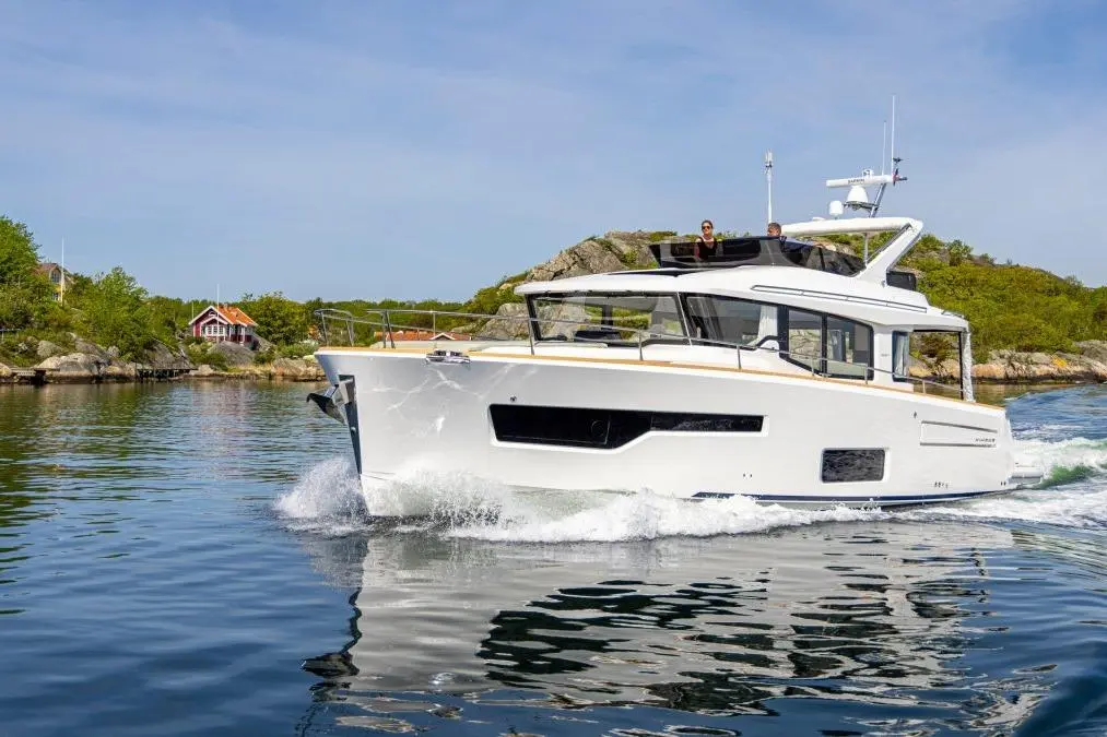 A luxurious motor yacht glides through calm waters, surrounded by greenery and a distant red house on a sunny day.