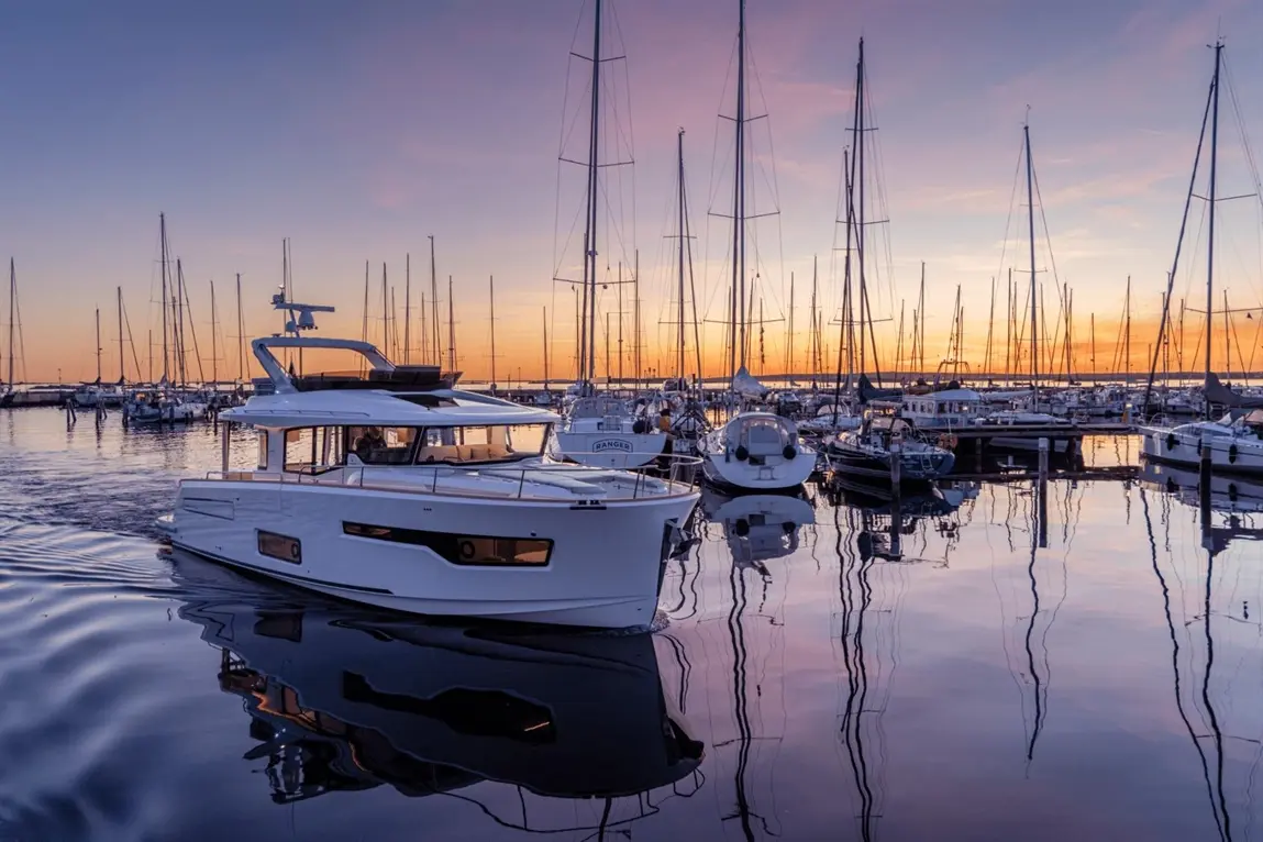 A sleek white motorboat navigates a marina at sunset, surrounded by numerous sailboats and tall masts reflecting in calm waters.