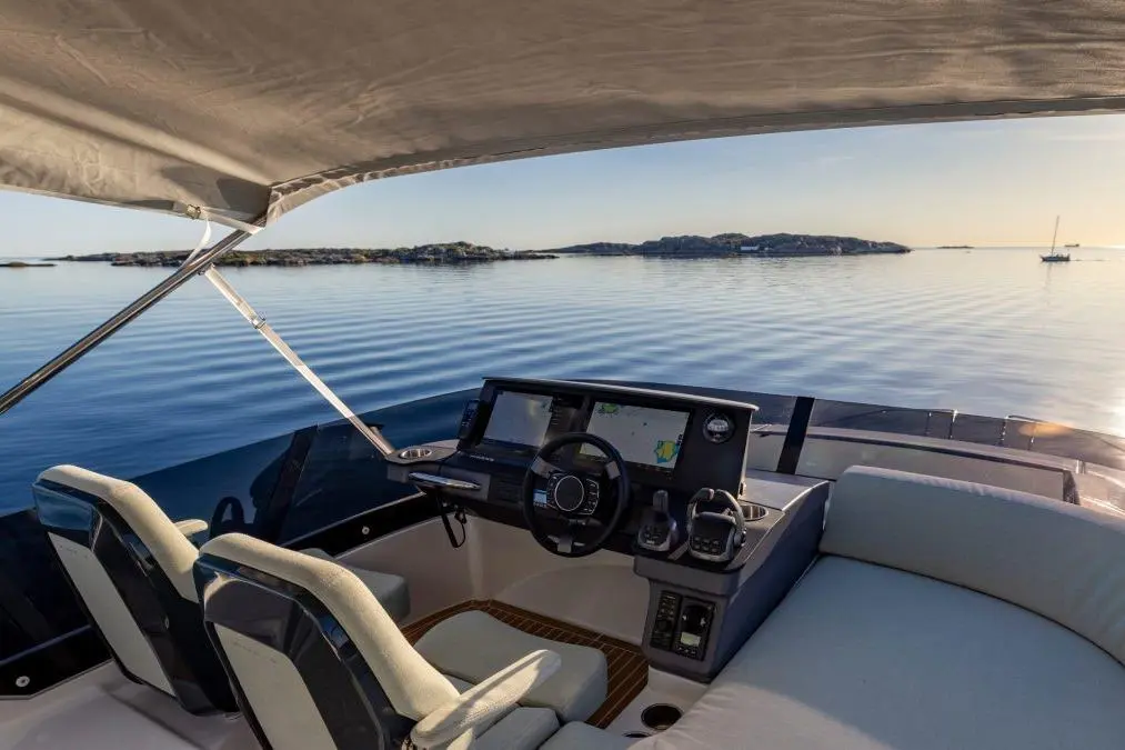View from the cabin of a boat, featuring a dashboard with navigation screens, two seating areas, and a calm water backdrop.
