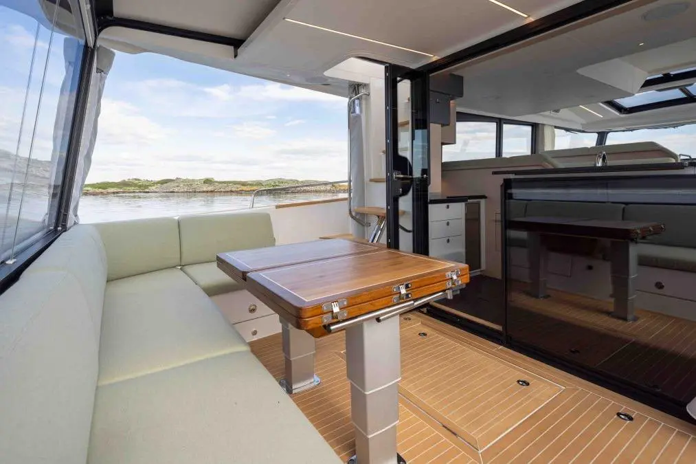 Interior of a modern boat featuring a wooden table, light upholstery, and large windows with a view of the water and landscape.