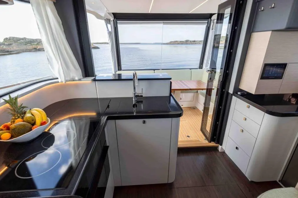 Modern yacht interior featuring a sleek black kitchen, fruit bowl, and glass doors opening to scenic waterfront views.