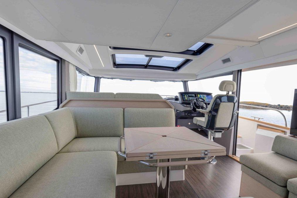 Modern boat interior featuring a plush seating area, large windows, control panel, and a central dining table.