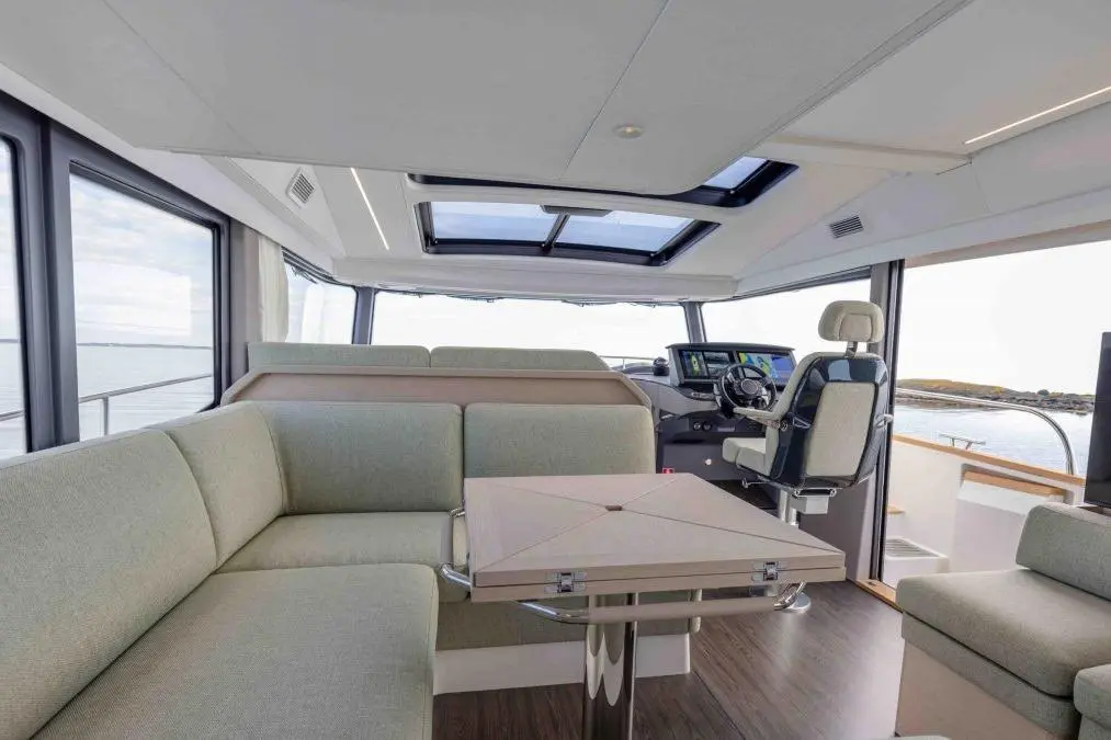 Modern boat interior featuring a plush seating area, large windows, control panel, and a central dining table.