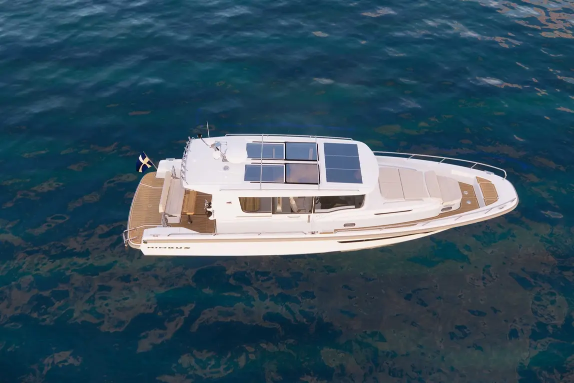 Aerial view of a sleek white boat with solar panels, anchored in clear blue water, showcasing a wooden deck and flag.