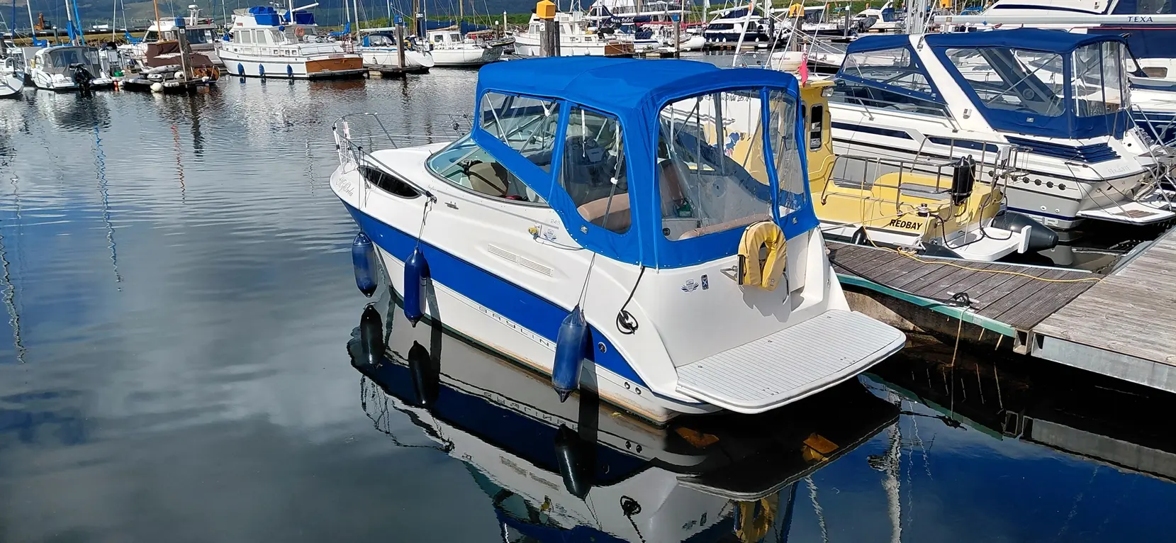 A boat with a blue canopy is docked in a marina surrounded by various other boats and reflections in the water.
