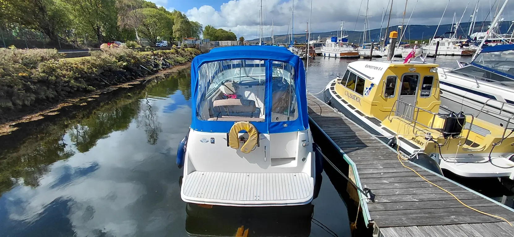 A boat with a blue canopy moored at a dock, reflecting in calm waters, alongside a yellow vessel. Green trees in the background.