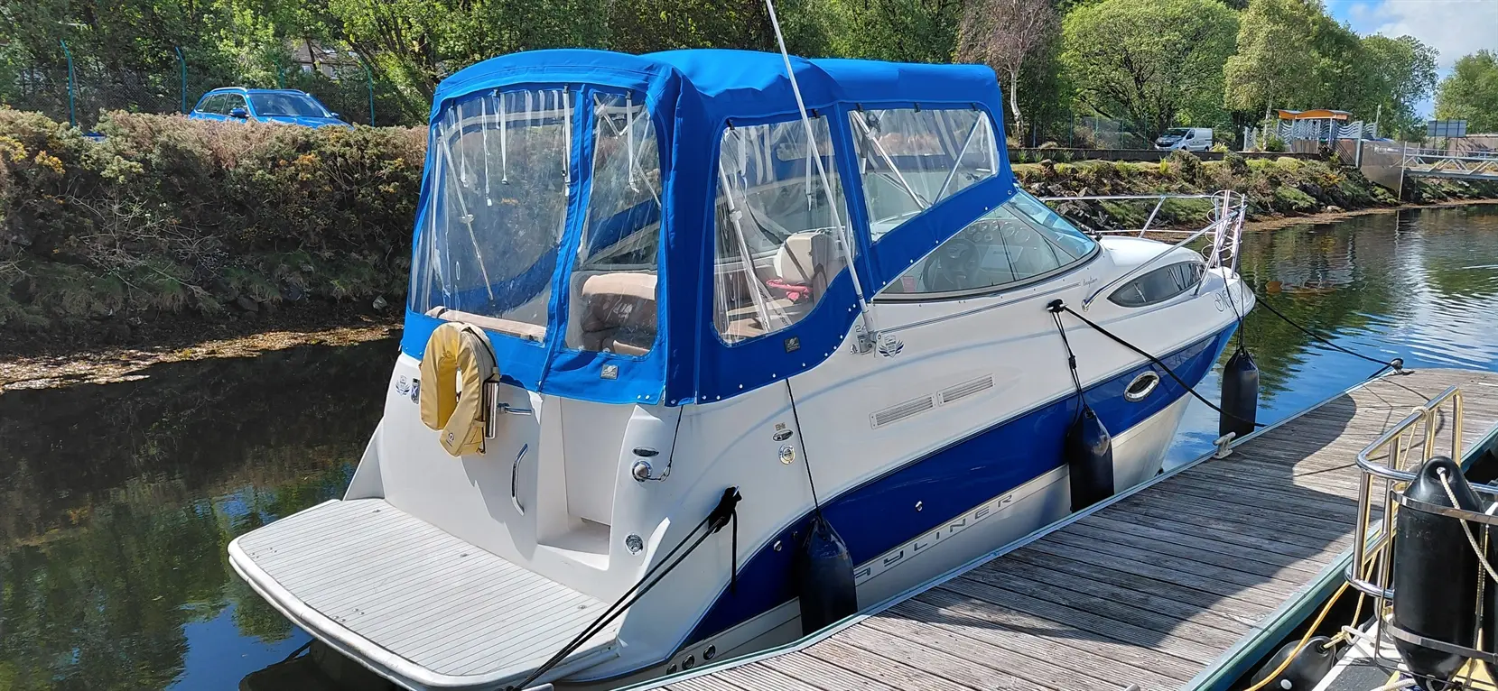 A blue and white cabin cruiser boat is docked with a life buoy, set against a sunny riverside backdrop.