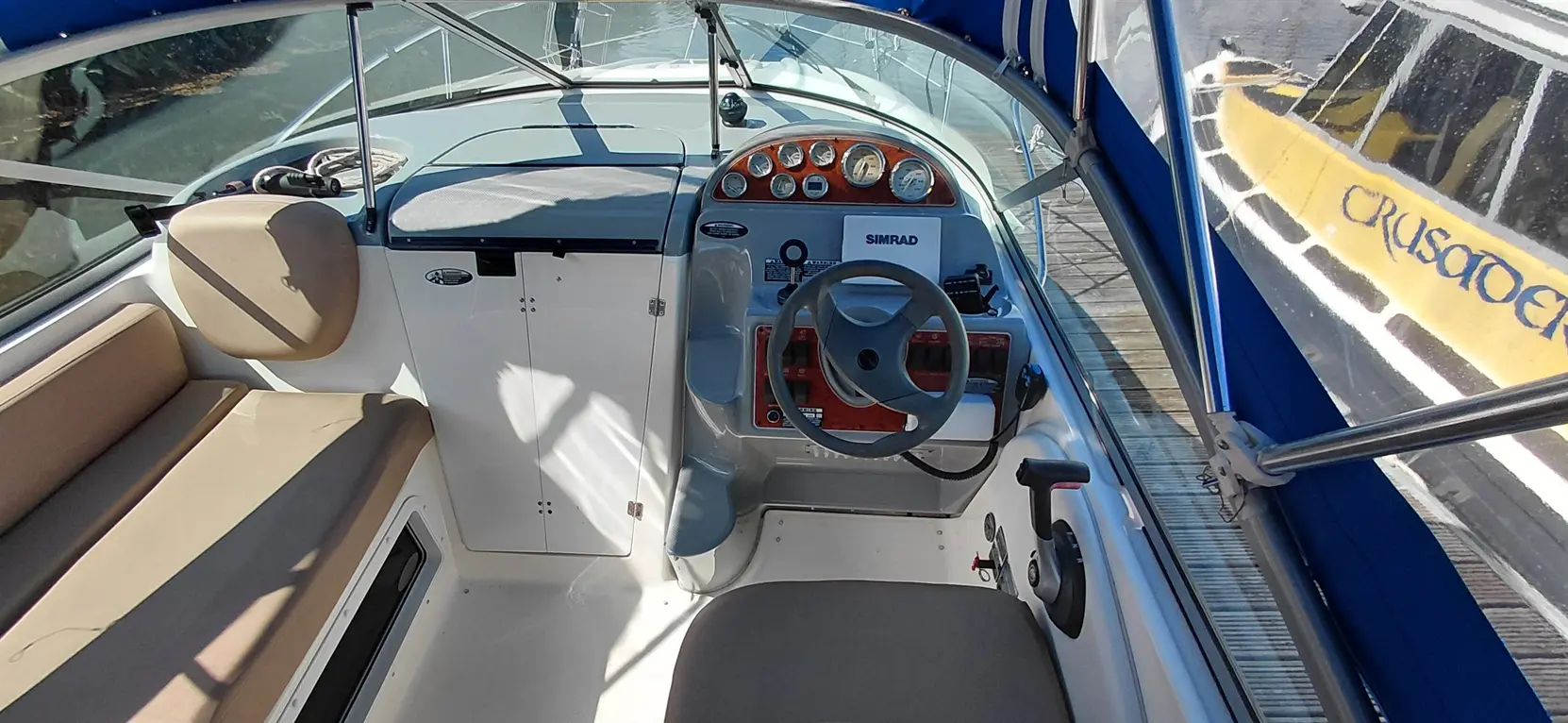 Interior of a boat showing the dashboard with gauges, steering wheel, and seats under a blue canopy. Visible text: "SIMRAD.