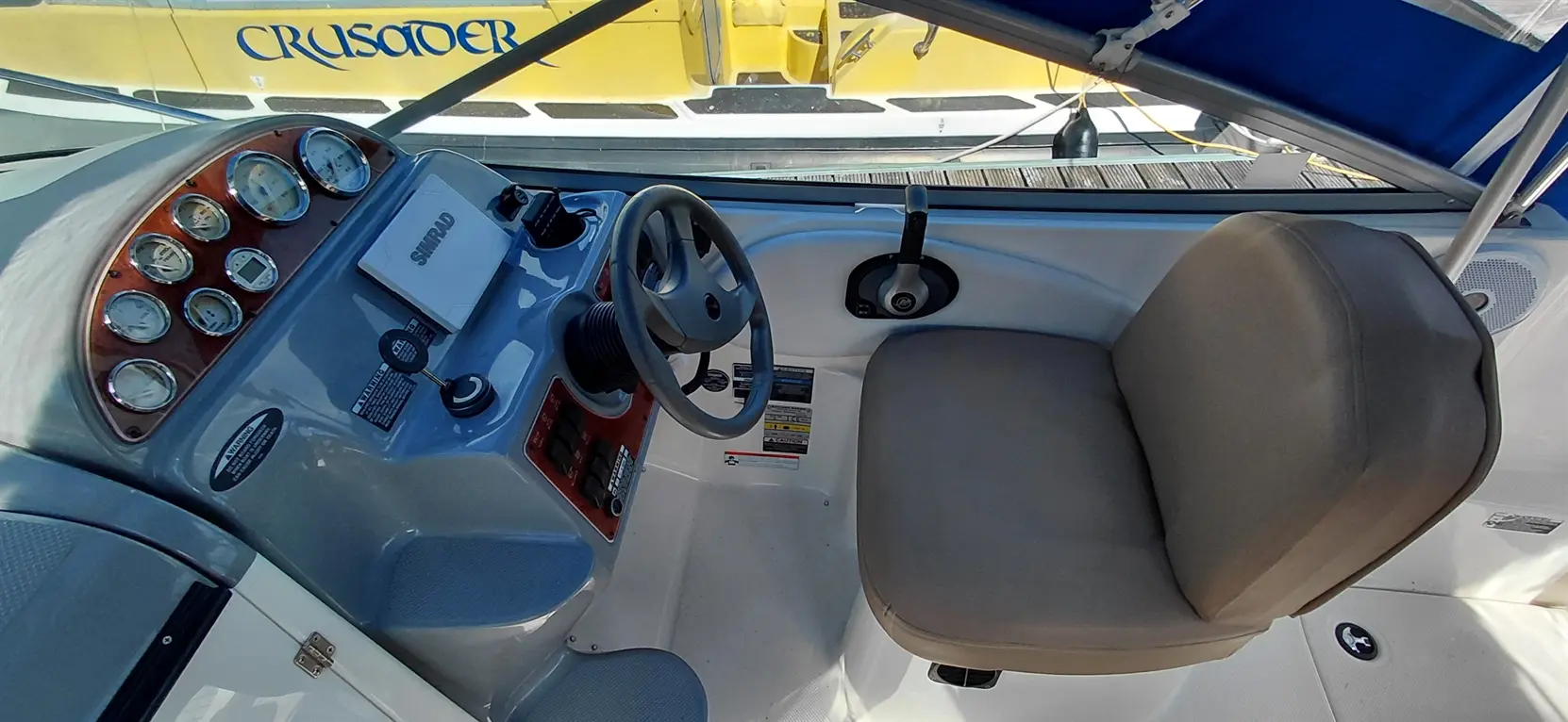 Boat cockpit featuring a control panel with gauges, a steering wheel, and a cushioned seat, overlooking a yellow boat labeled "CRUSADER.
