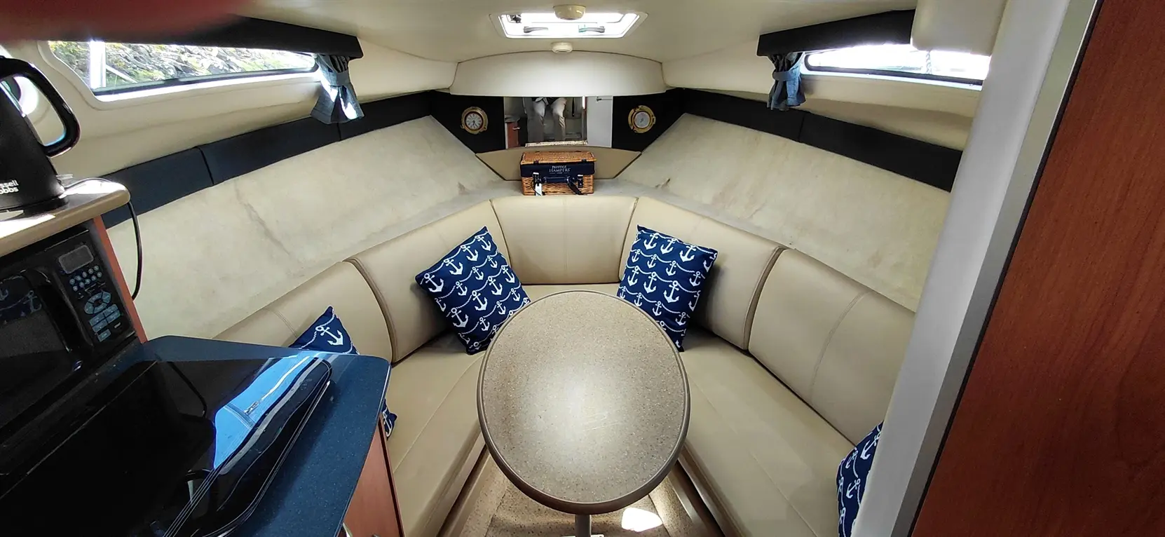 Interior of a boat featuring a light-colored seating area with anchor-patterned pillows, a round table, and a kitchenette.