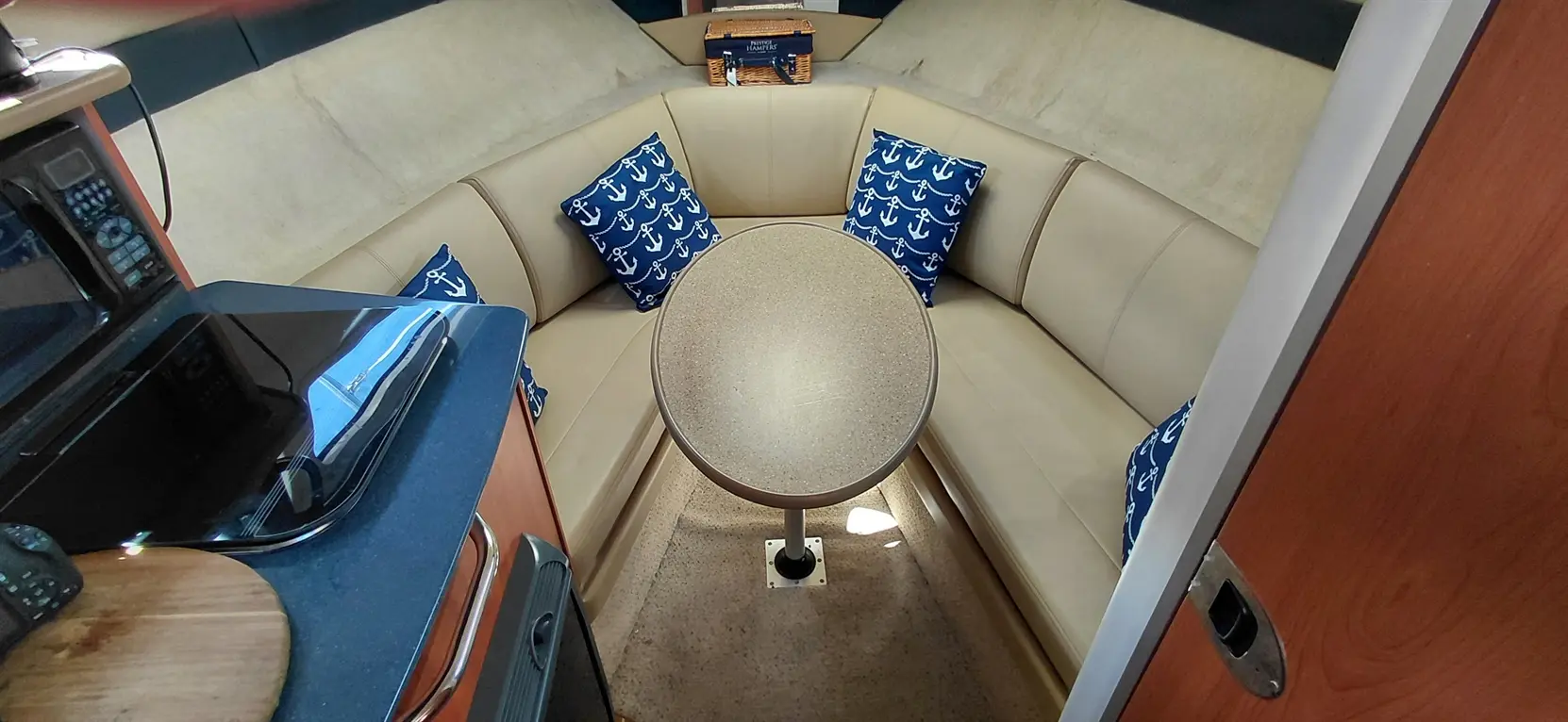 A cozy boat interior featuring beige seating with anchor-patterned pillows, a round table, and a compact kitchen area.