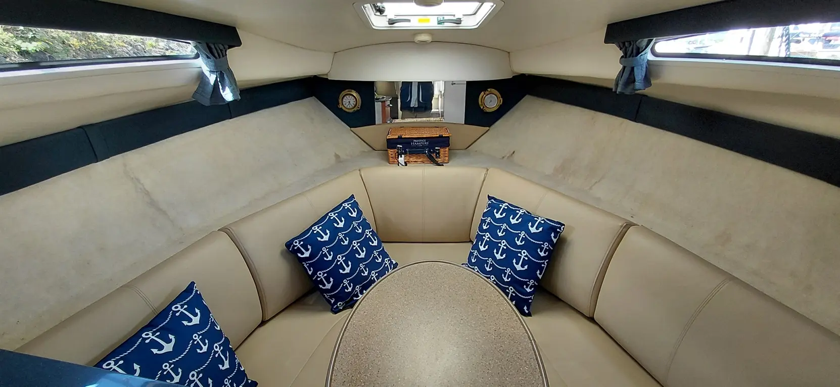 Cozy yacht interior with beige seating, blue anchor-patterned pillows, a round table, and decorative clock; natural light enters through windows.