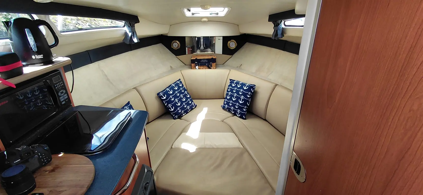 Cozy interior of a boat featuring a curved sofa with anchor-patterned cushions, kitchenette, and large windows for natural light.