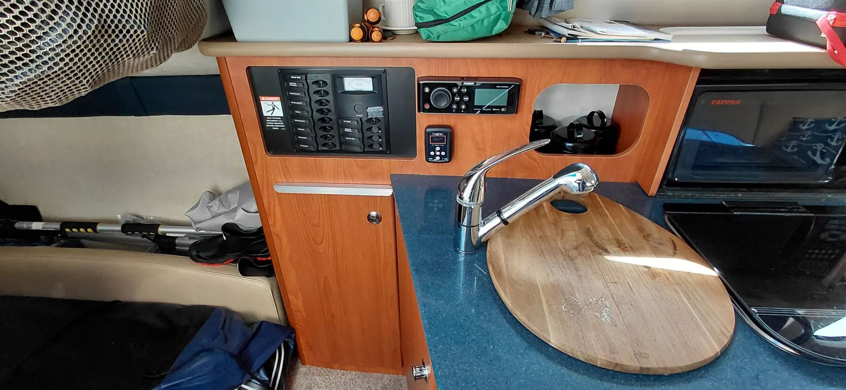 Compact boat kitchen with a sink, wooden cutting board, radio, electrical panel, and storage area.