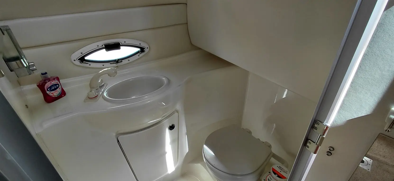 Compact marine bathroom with a round sink, toilet, Carex hand soap, and a small porthole for natural light.