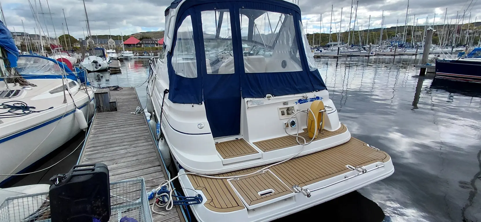 A motorboat with a blue canopy is docked at a marina surrounded by other boats and a calm water reflection.