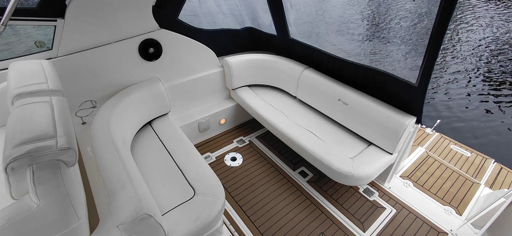 Interior of a boat featuring white vinyl seating and wooden flooring, with a view of water outside through window.
