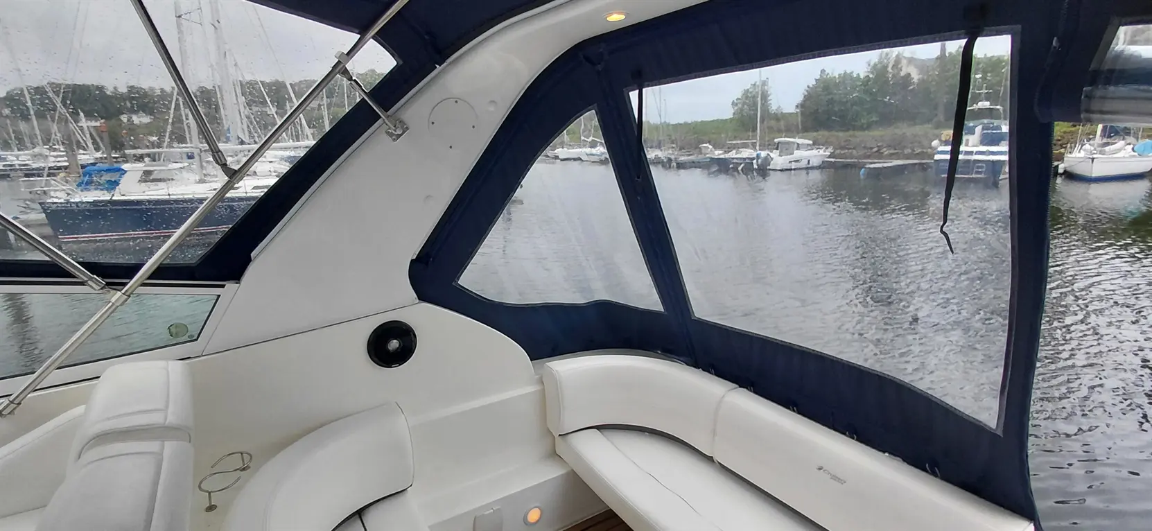 Interior view of a boat with white seating, rain on the windows, and a waterfront filled with parked boats.