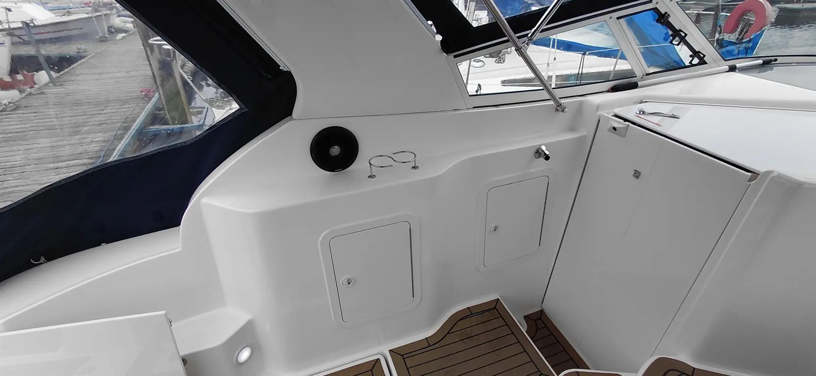 Interior view of a boat showing white seating, a speaker, storage compartments, and a wooden floor with boat mooring in the background.