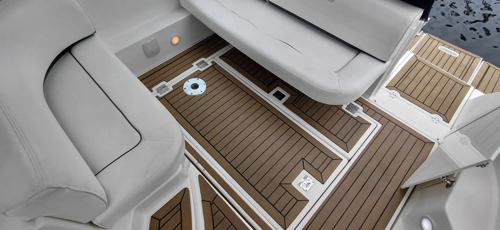 Interior of a boat showing a light-colored seating area and a wooden deck with detailed textures and a flush-mounted hatch.