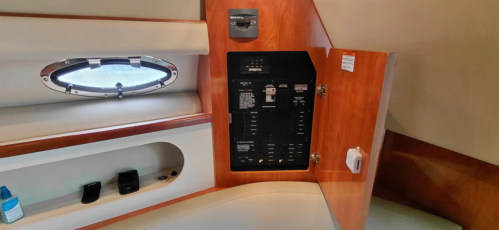 Interior of a boat showing a circuit breaker panel, porthole window, and storage area with various items.