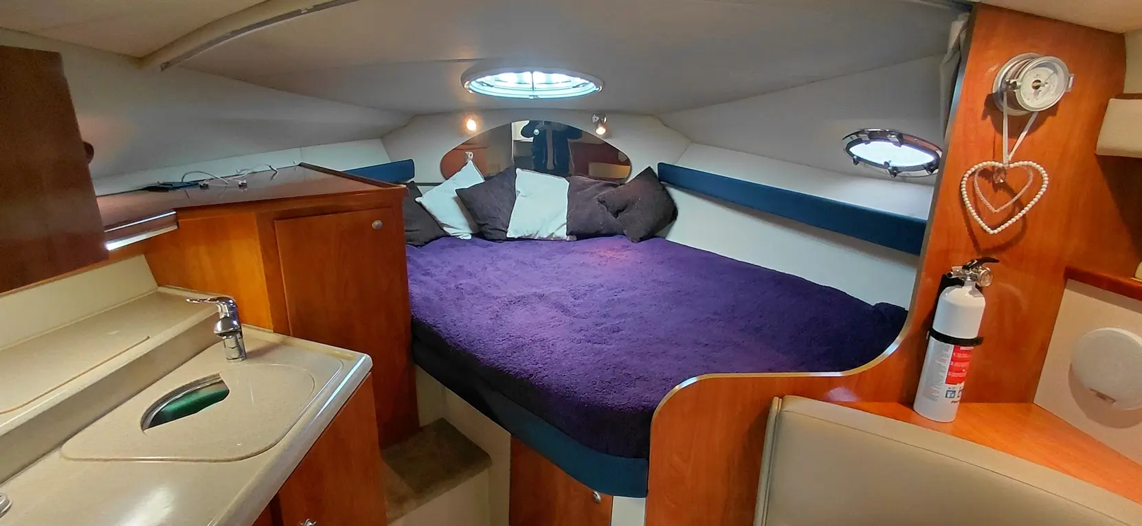 Interior of a boat showing a cozy sleeping area with a purple blanket, pillows, a small sink, and a fire extinguisher.