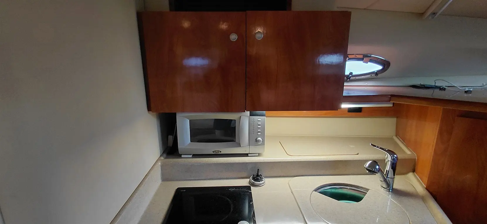 Compact marine kitchen featuring microwave, sink, and countertop with a stovetop; includes wooden cabinets and a porthole.