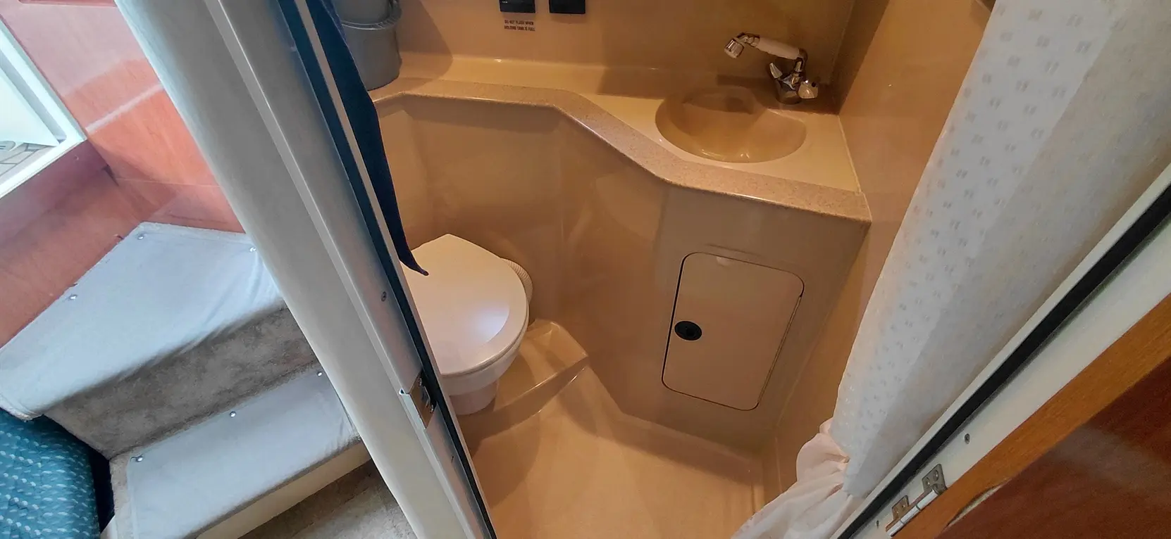 Compact boat bathroom featuring a toilet, small sink, and storage access panel, with steps visible outside.