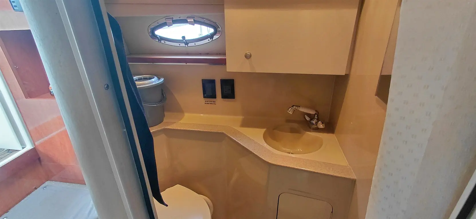 Compact boat bathroom featuring a toilet, sink, and window, with a trash bin and wall-mounted controls visible.