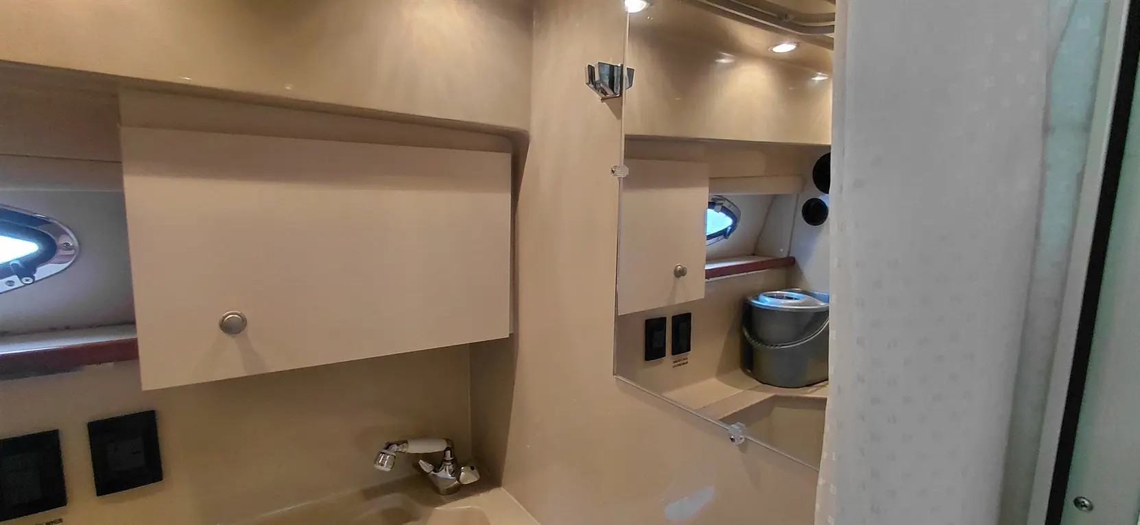 Interior of a boat bathroom with cabinets, a mirror, a sink, and a window, featuring a silver bucket and light fixtures.