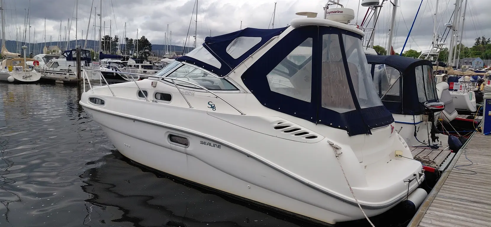 White Sealine S8 boat docked at a marina, surrounded by other vessels and masts, under a cloudy sky.