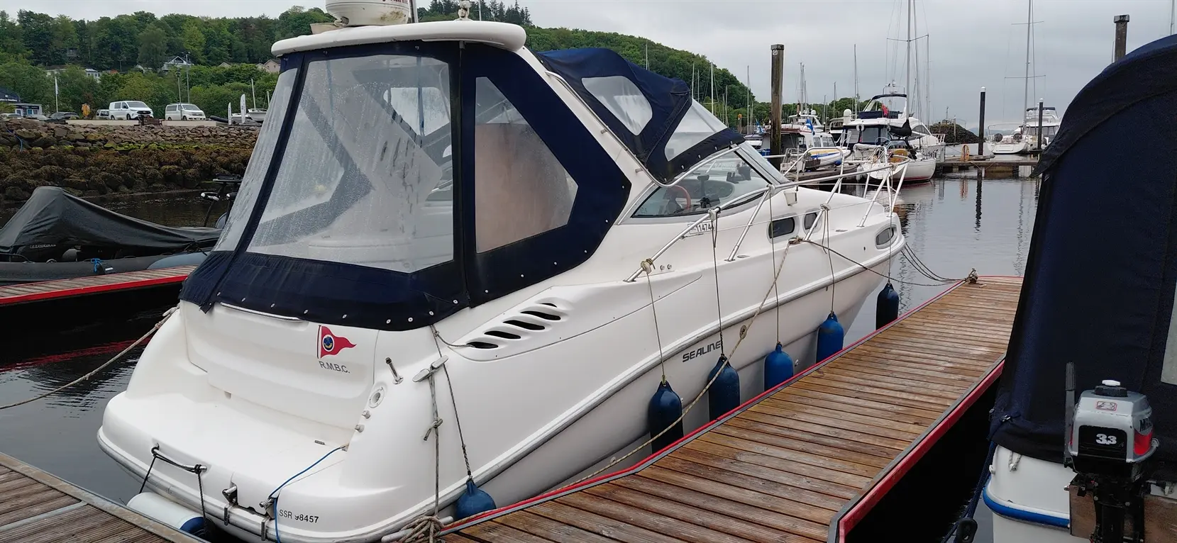 A white Sealine boat with a navy cover is docked beside wooden piers in a marina, surrounded by other boats and greenery.