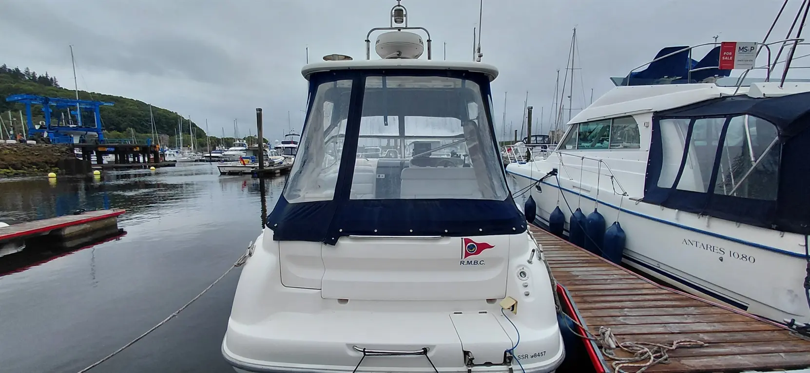 A moored motorboat with a blue canopy in a marina, surrounded by other boats and a boat lift in the background.