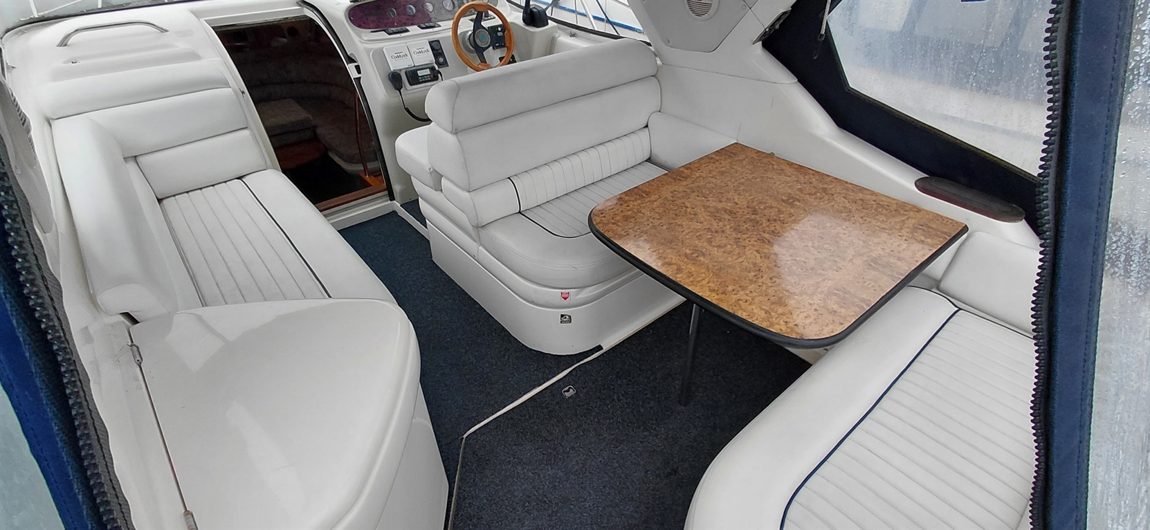 Interior of a boat featuring white leather seating, a wooden table, and a blue carpet with a view of the dashboard.